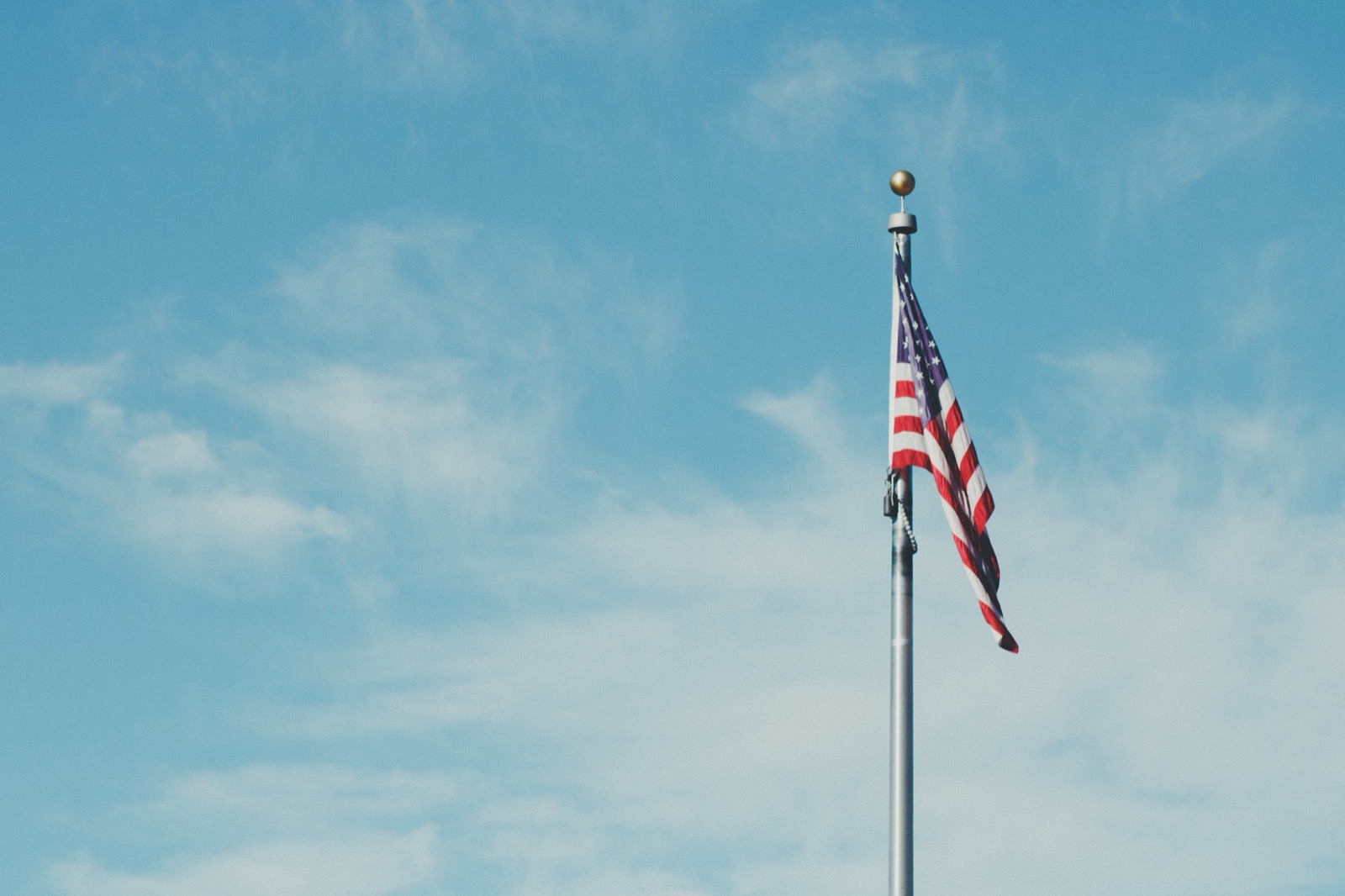A United States flag flying from a tall pole against a soft, pale-blue sky.